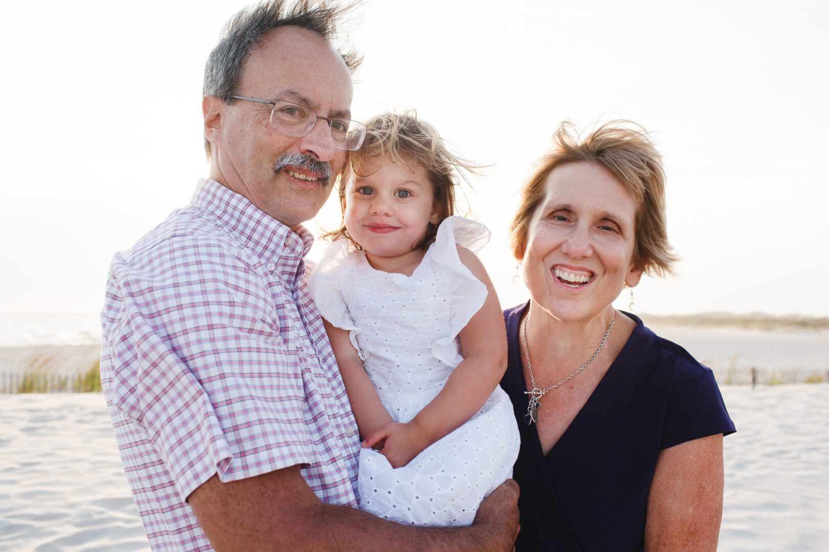 On a sandy Cape May beach, a man and woman smile warmly as the man holds their young child in a white dress, capturing precious family photos.