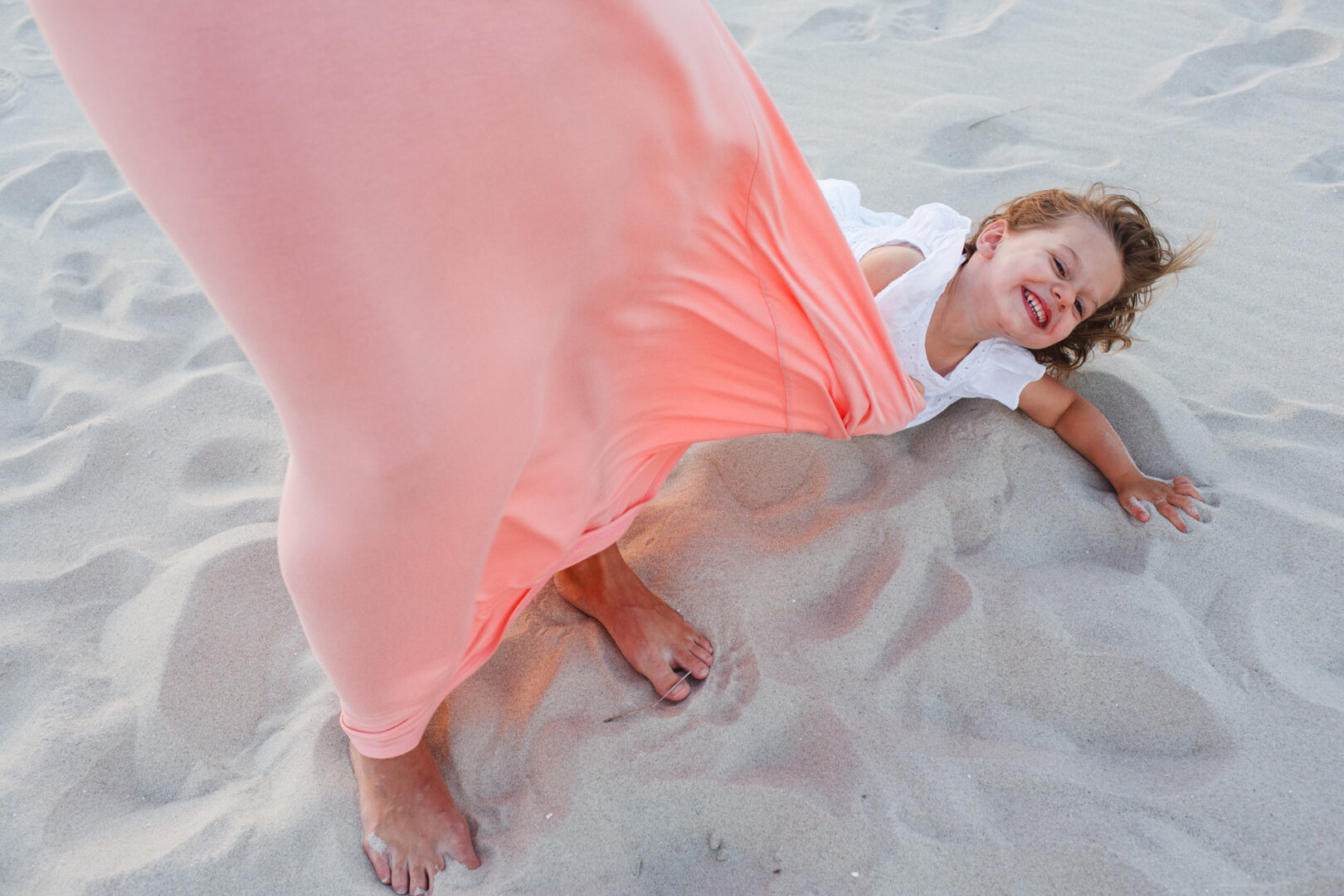 A child laughing while lying on the sand, captured in Cape May family photos. An adult's legs, wearing a flowing peach skirt, stand nearby.