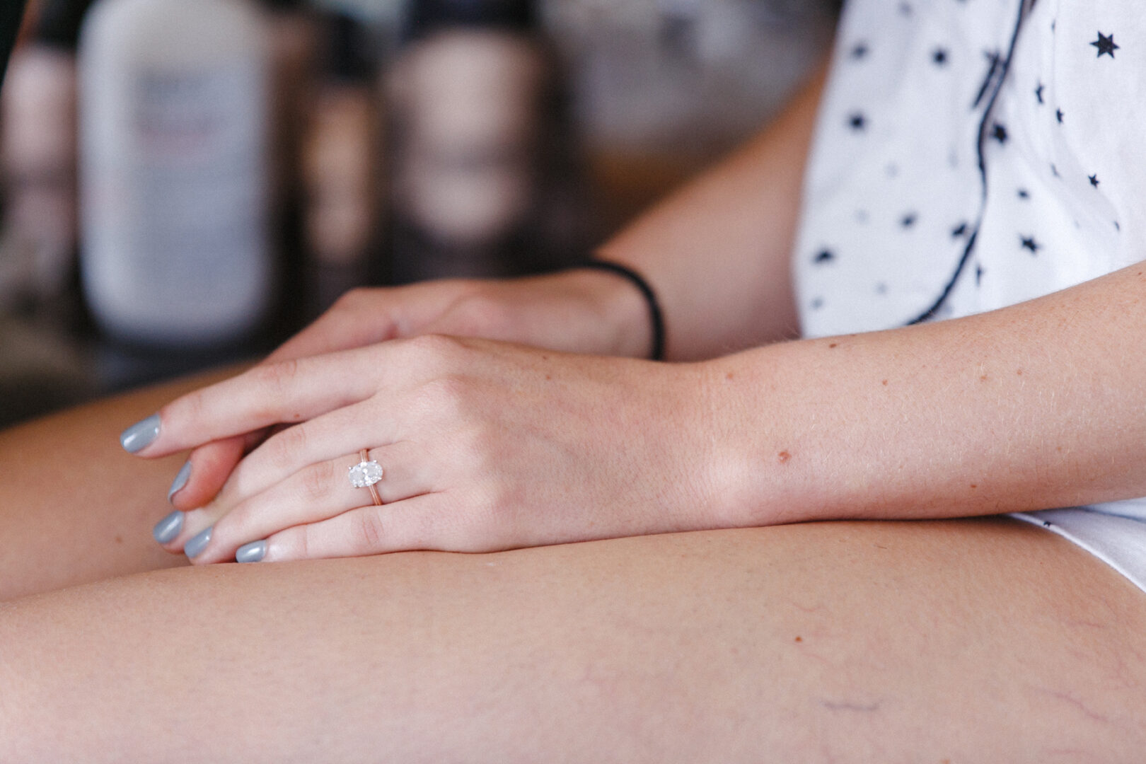 A close-up captures a person in a white shirt adorned with star patterns and shorts, showcasing a ring, hands gracefully resting in their lap. Gray nail polish complements the look. The blurred background suggests an elegant setting, reminiscent of a Franklin Institute wedding's timeless charm.