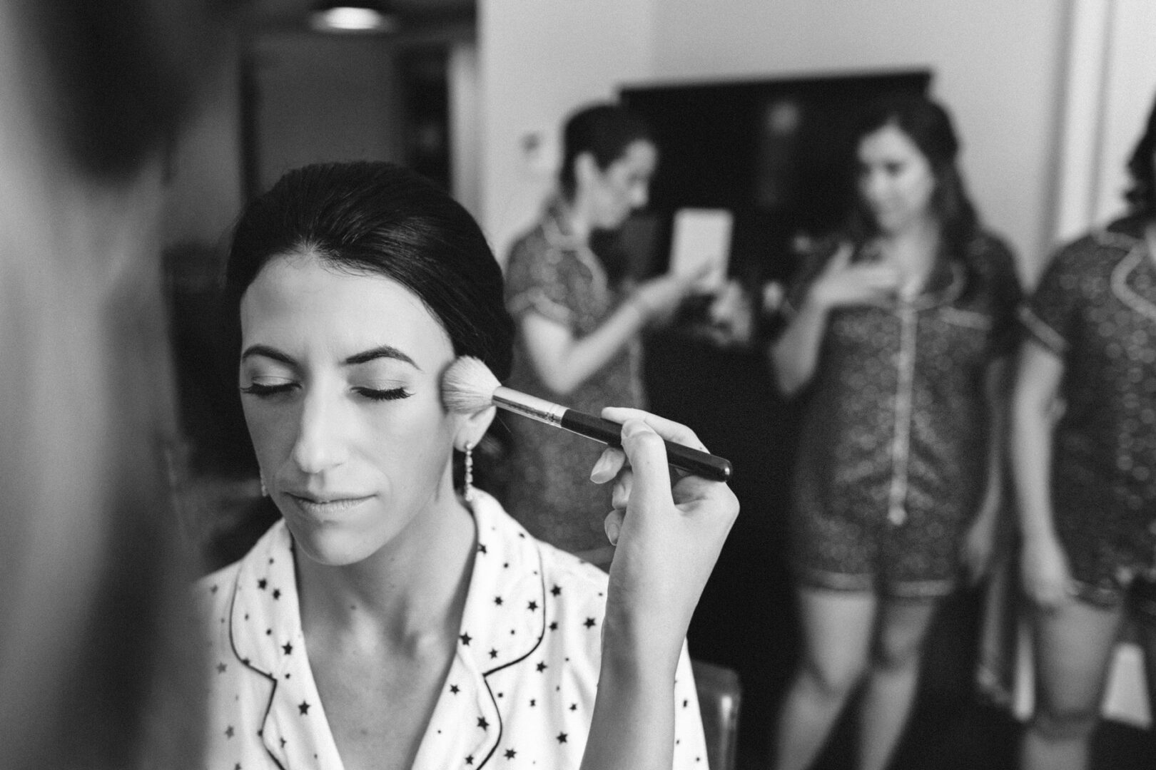 At a Franklin Institute wedding, a woman gets makeup applied with a brush, eyes closed, as two others in matching outfits chat in the background.