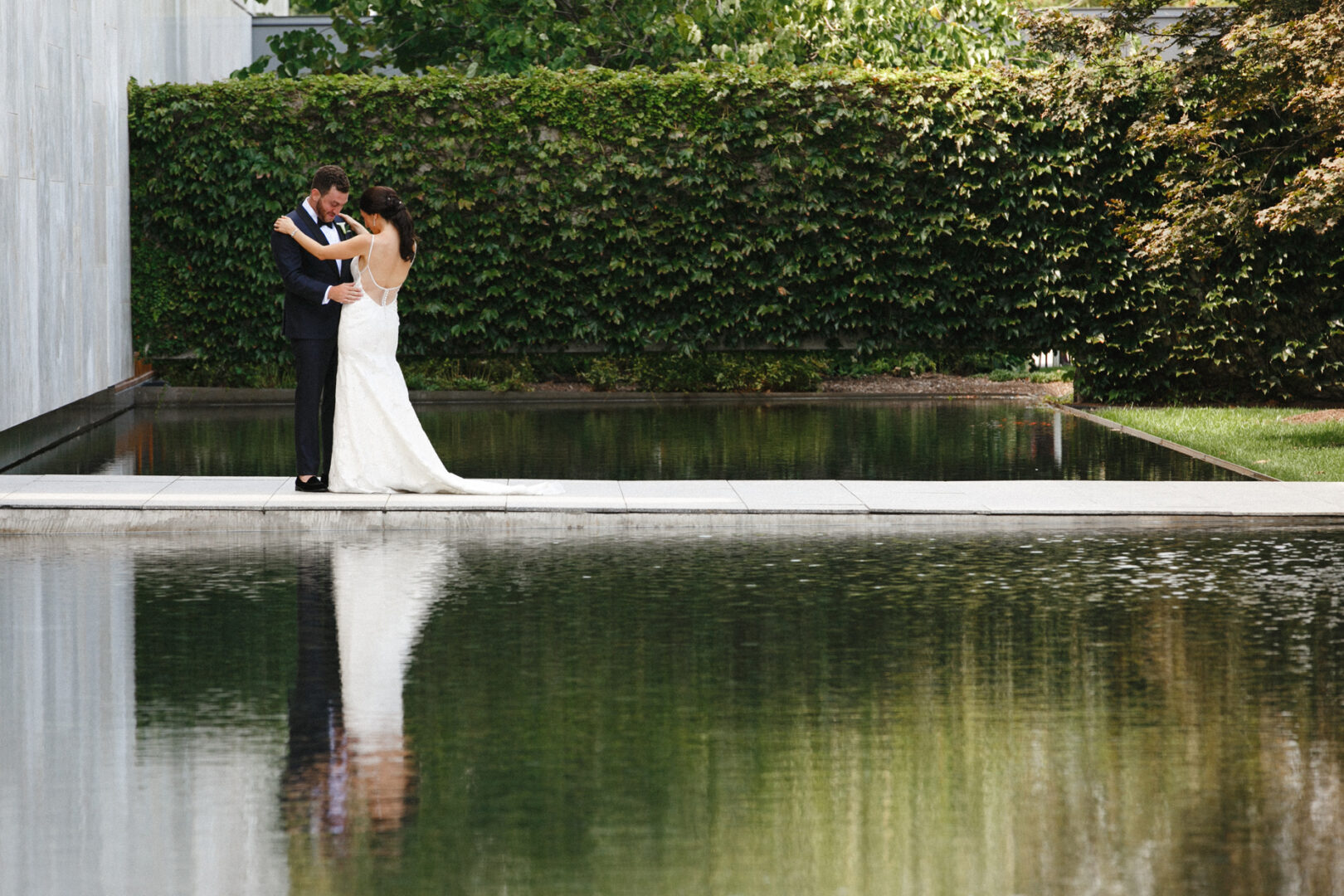 A couple, dressed in formal wedding attire, stands embracing near a reflective pool at the Franklin Institute Wedding venue, with lush greenery providing a stunning backdrop.