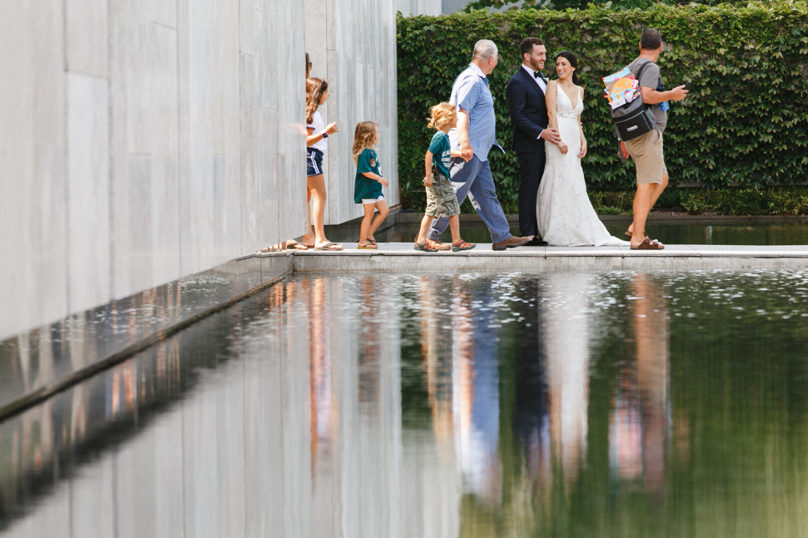 At a Franklin Institute wedding, a bride and groom stroll with their guests by a reflective pool, lush greenery providing a serene backdrop.