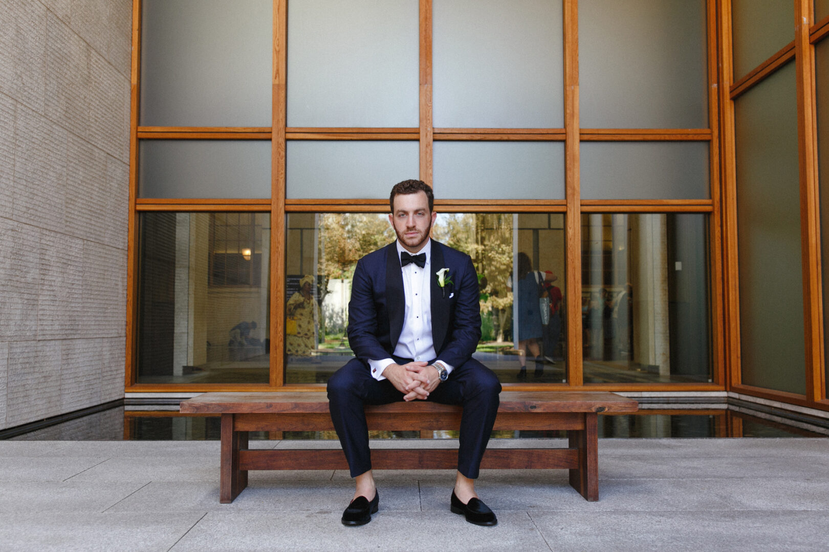 A man in a suit sits on a wooden bench, framed perfectly by the grand windowed backdrop of the Franklin Institute, where countless dreams come alive during enchanting weddings.