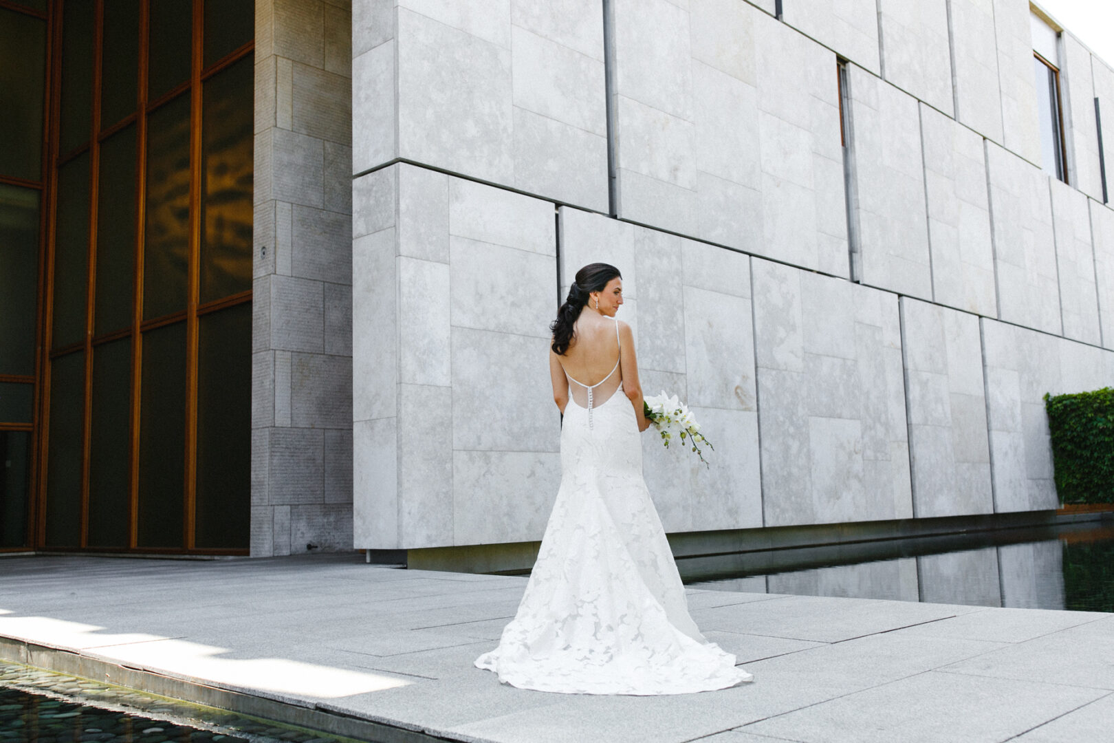 A bride in a white wedding dress stands on a stone platform by the water, facing away from the camera, with the iconic backdrop of the Franklin Institute setting a modern tone for her wedding.