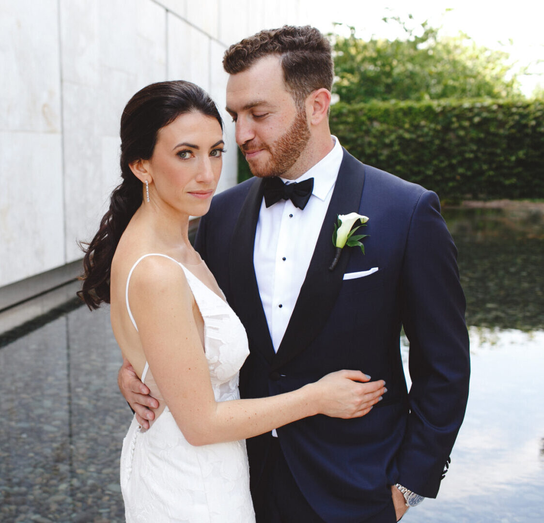 Bride and groom pose together by a reflective pool at their Franklin Institute wedding, with the groom looking lovingly at the bride. She wears a white gown; he dons a black tuxedo with a white boutonniere.