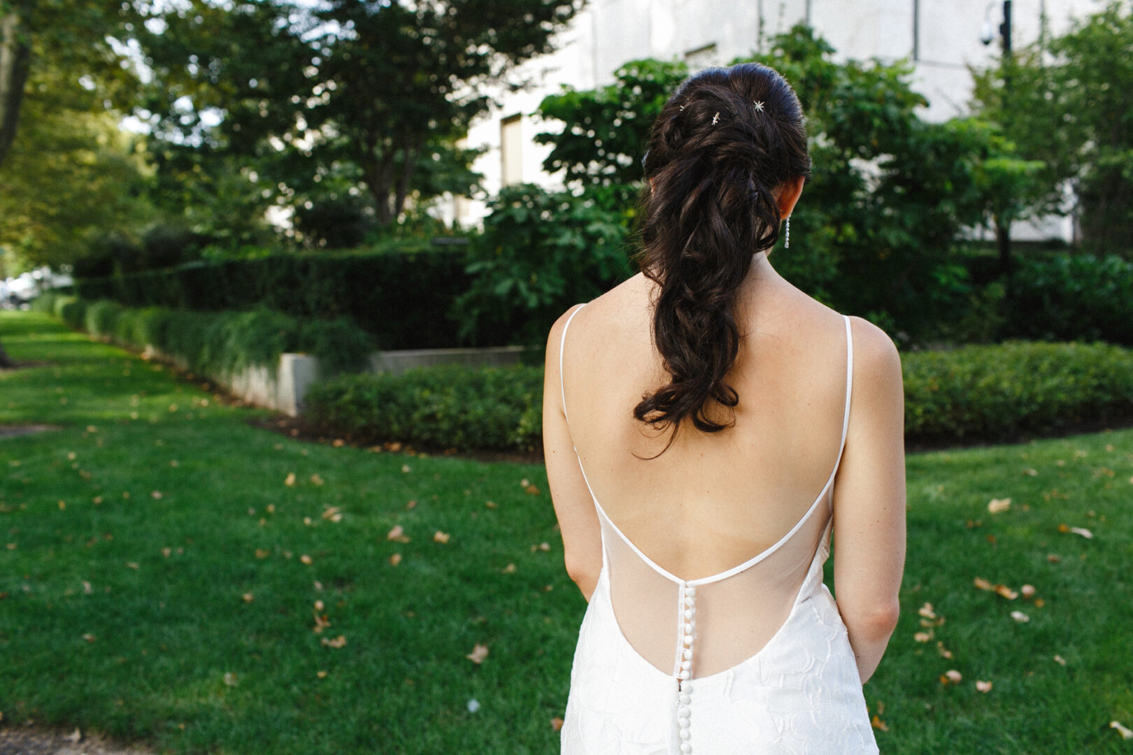 A woman in a wedding dress with an open back stands on the grass at a Franklin Institute wedding, facing away, her hair in a loose braid with lush greenery in the background.