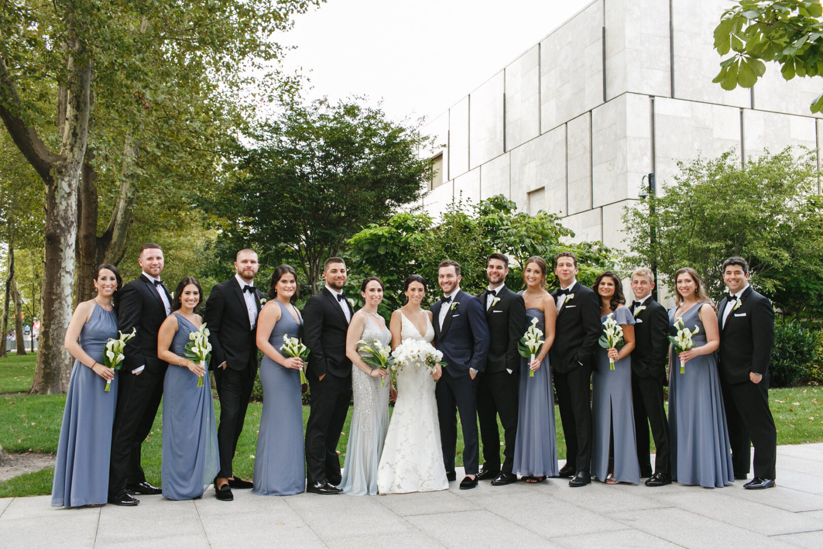 A wedding party poses outdoors at a Franklin Institute Wedding, with the bride and groom in the center, surrounded by bridesmaids in blue dresses and groomsmen in black suits. Trees and a building form the beautiful backdrop.