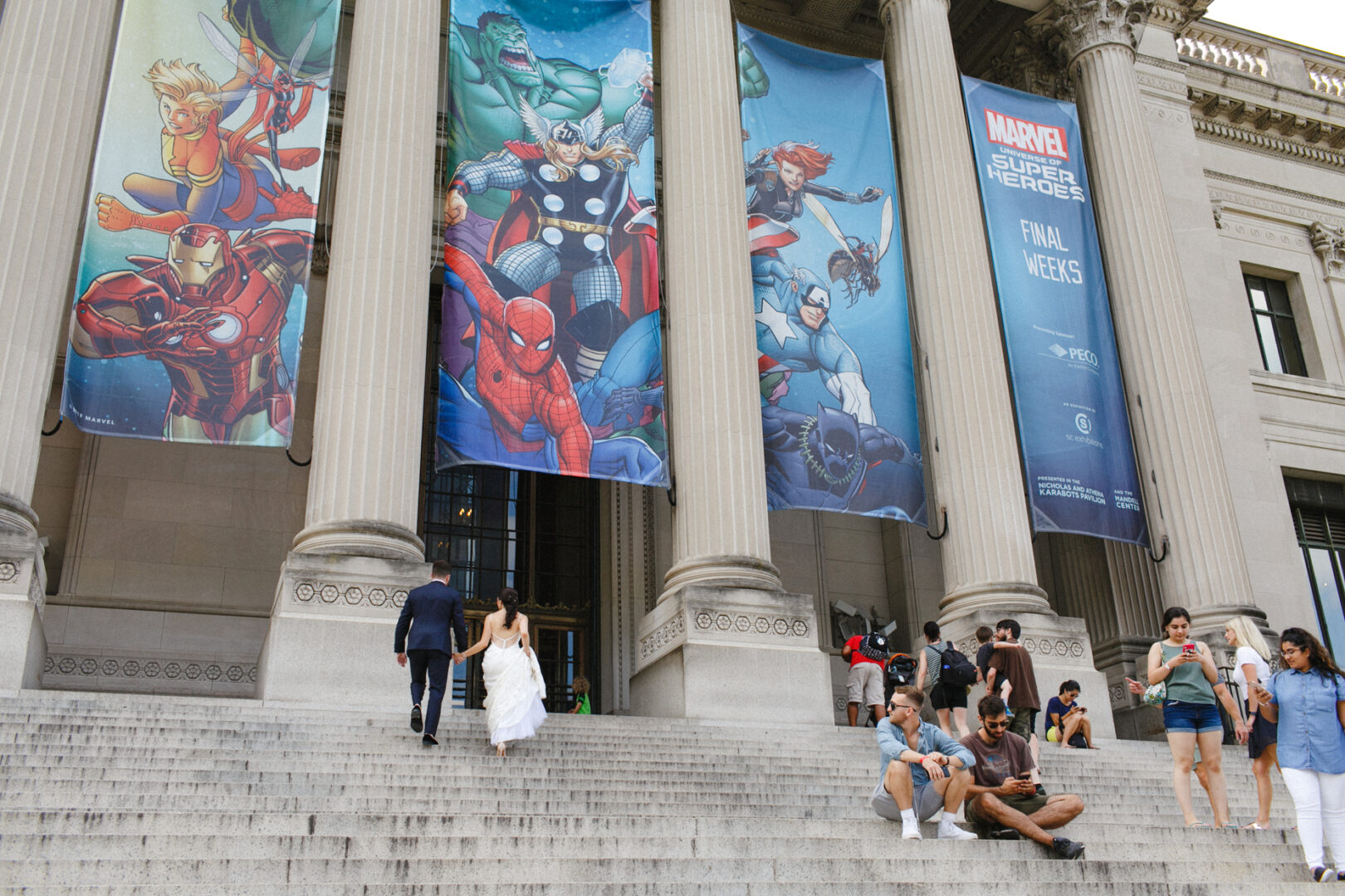 Steps leading to a building adorned with large superhero banners evoke the excitement of a Franklin Institute Wedding. People are walking or sitting on the steps, buzzing with anticipation as they discuss the upcoming celebration within this iconic venue.