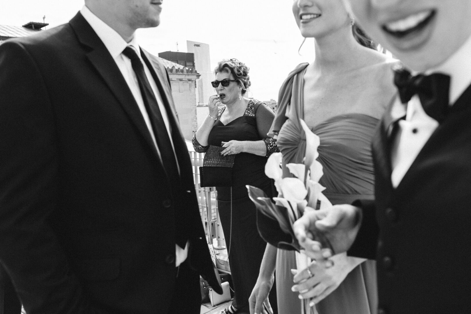 A group of people in formal attire, two men in suits and a woman holding flowers, stand and converse outdoors, reminiscent of a Franklin Institute wedding. A woman in sunglasses lingers in the background. Black and white photo.