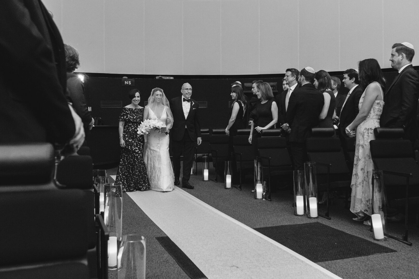 At the Franklin Institute, the bride gracefully walks down the aisle with two escorts in a formal setting, surrounded by standing guests.