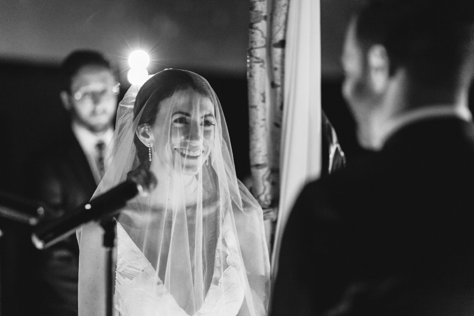 A bride smiles under a veil while facing the groom during a Franklin Institute wedding ceremony, with an officiant in the background. The scene is in black and white.