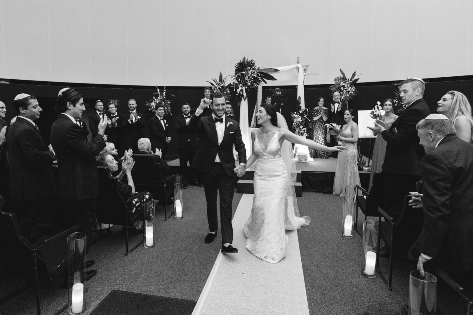 The bride and groom walk down the aisle hand in hand, smiling, as guests applaud beneath the stunning arches of the Franklin Institute Wedding venue.