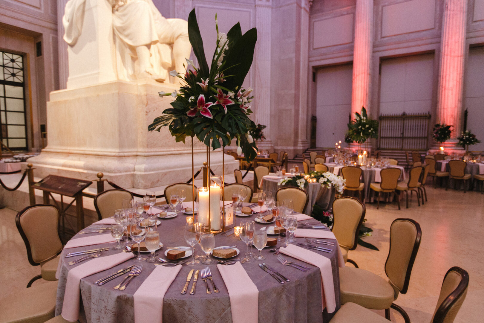Elegant table setting with candles and a floral centerpiece in a grand room at the Franklin Institute, complete with a large statue, stately columns, and soft pink uplighting.