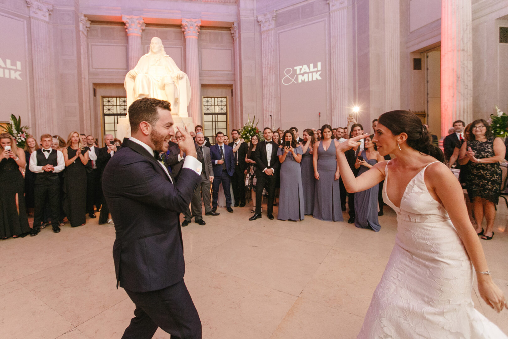 A couple dances joyfully in formal attire at a wedding reception, surrounded by guests in a grand hall at the Franklin Institute, with a large statue and "Tali & Mik" elegantly projected on the wall.