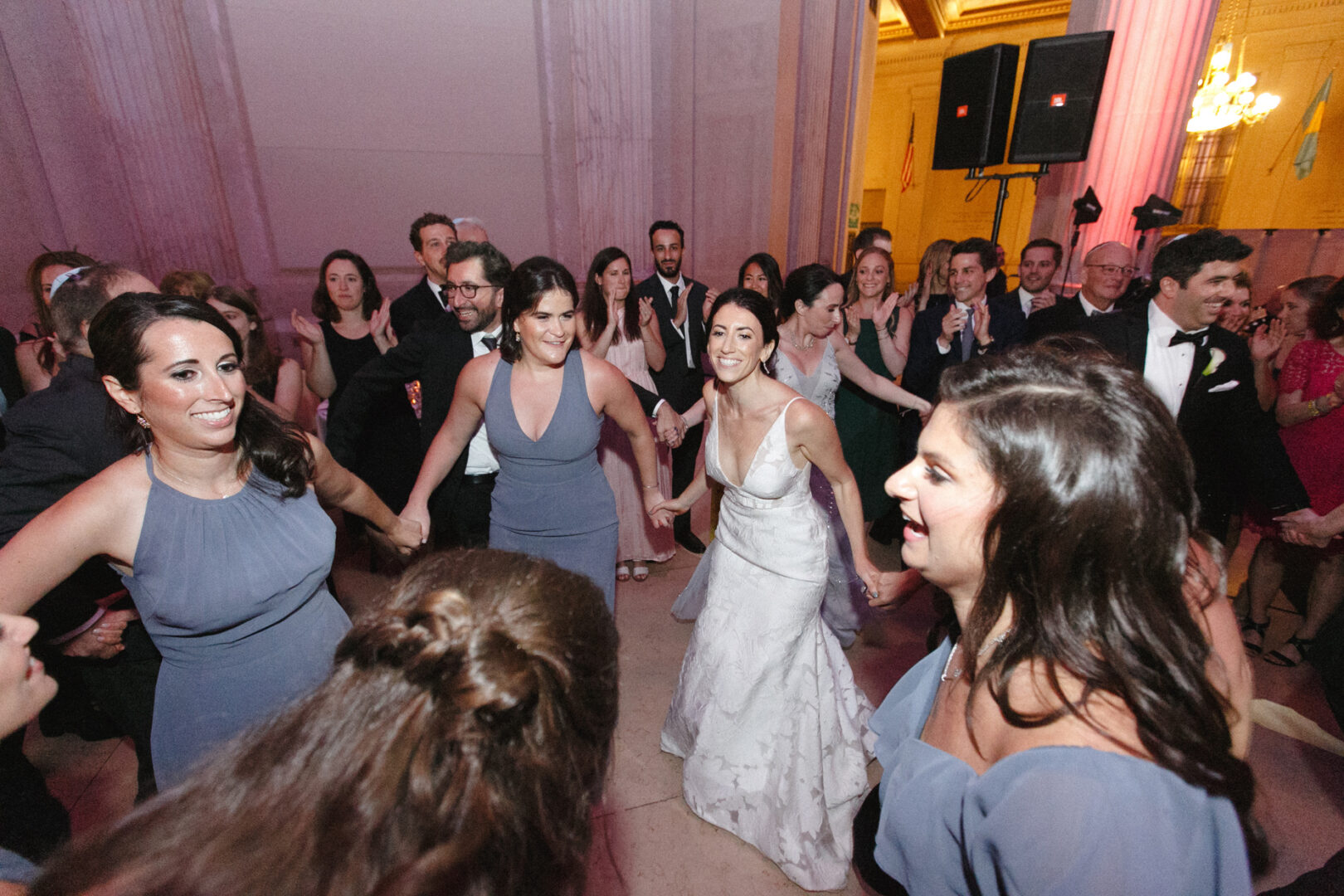 At a Franklin Institute wedding, a bride dances gracefully in a circle of people at an indoor event. Guests in formal attire gather around, clapping and smiling, celebrating the joyous occasion.
