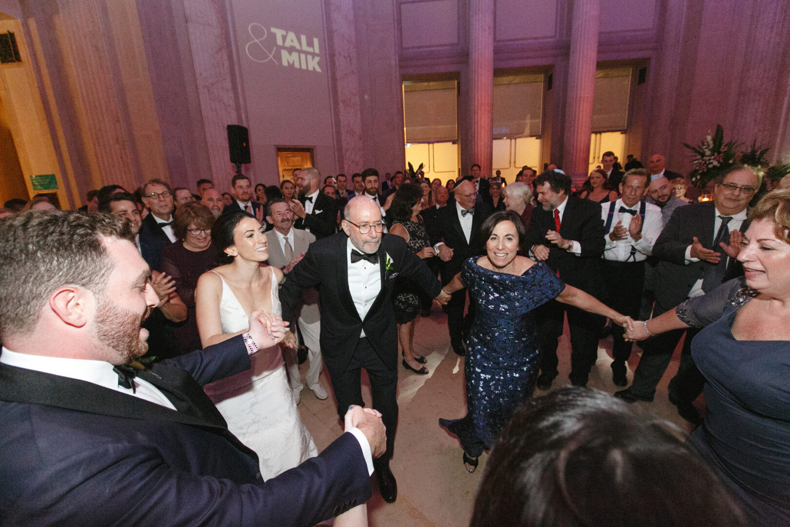 A group of people, dressed in formal attire, dance joyfully in a circle at an indoor Franklin Institute wedding, surrounded by tall columns and soft lighting.
