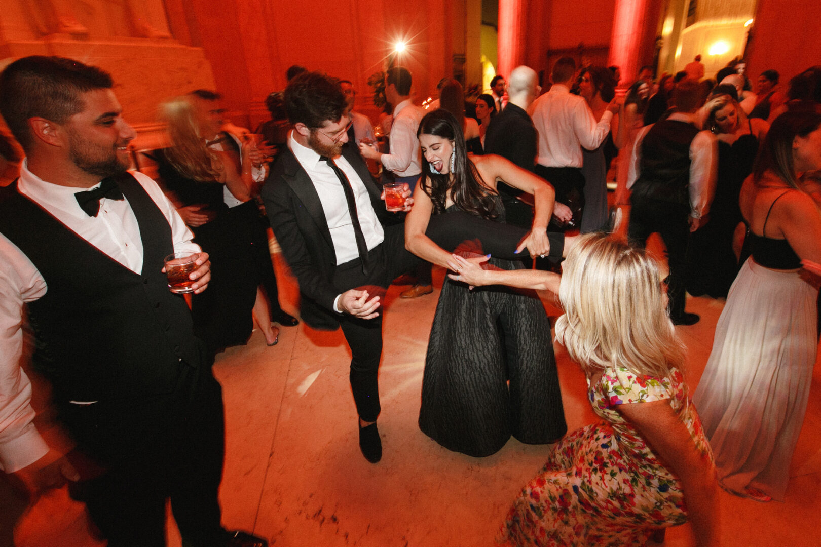 At a Franklin Institute wedding, people dance energetically at a lively indoor event, with some holding drinks. The warmly lit room is filled with guests in formal attire, celebrating the joyous occasion in style.