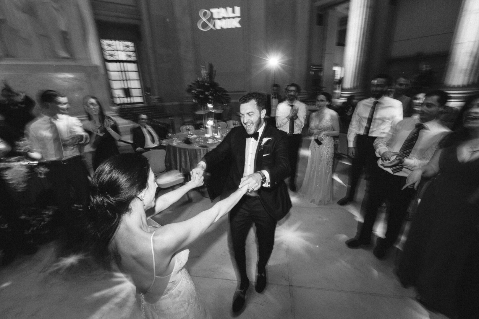 A couple dances gracefully in the center of a grand room at the Franklin Institute, surrounded by smiling and clapping guests. The man dons a suit while the woman twirls in her elegant dress. A projection on the wall reads "Tali & Nick.