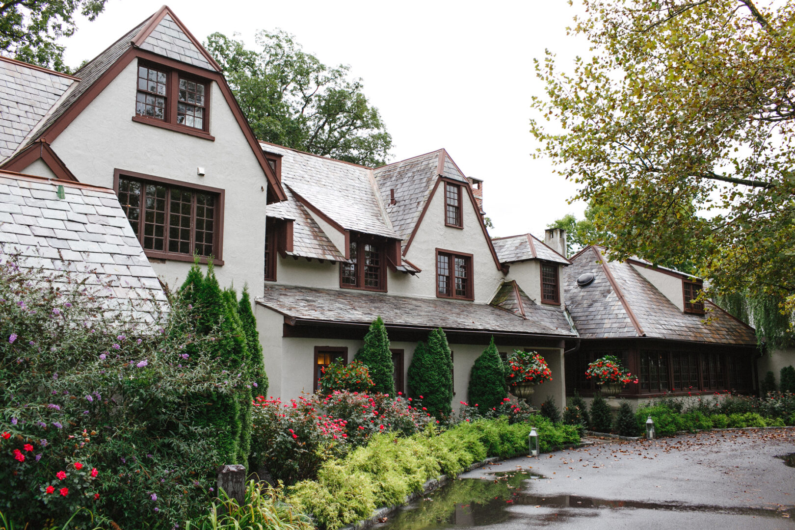 A charming venue reminiscent of a large house with gabled roofs, Hotel Du Village is surrounded by lush greenery and flowered shrubs, creating the perfect backdrop for a romantic wedding under an overcast sky.