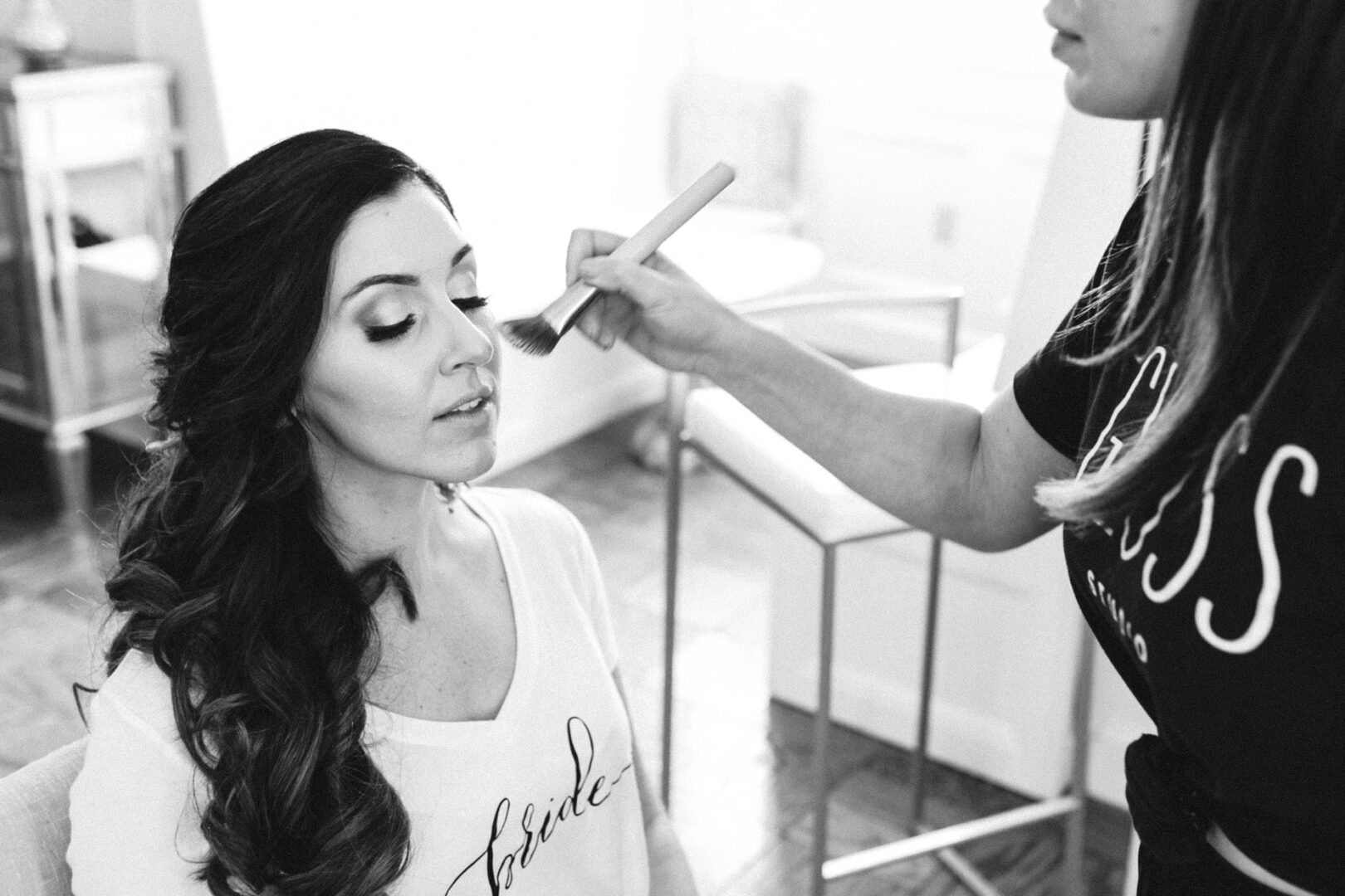 In a sunlit room at Hotel Du Village, a woman with long hair sits gracefully as another applies makeup with a brush. Her shirt proudly displays the word "bride," capturing the joyful anticipation of her wedding day.