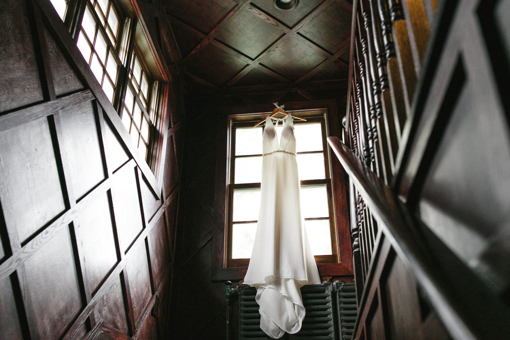A white wedding dress hangs in front of a tall window inside a wood-paneled room at Hotel Du Village, with stairs visible on the right.