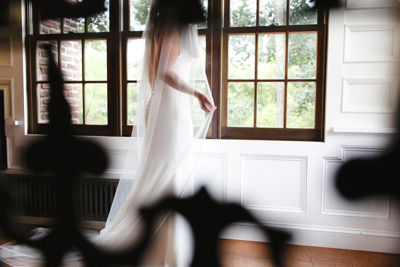 The bride, in a white dress and veil, stands gracefully near the large windows of Hotel Du Village, partially obscured by the decorative railing.