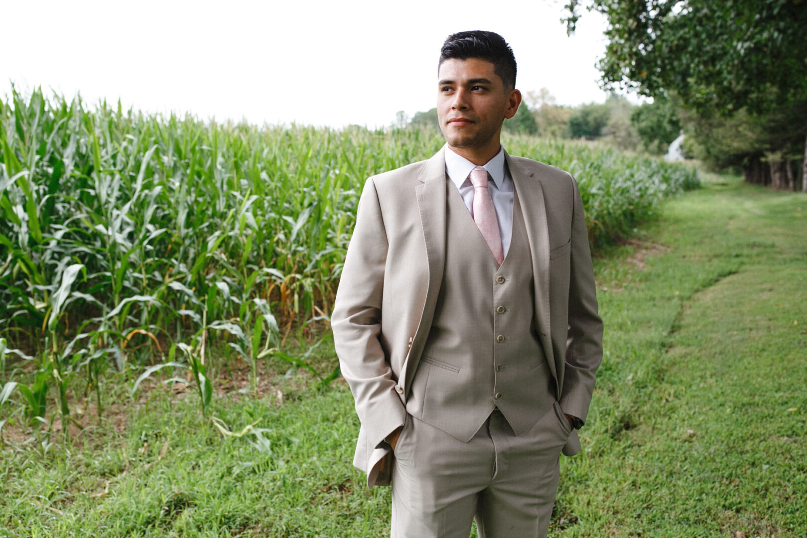A man in a beige suit and pink tie stands near a cornfield, with hands in pockets, looking ahead, as if envisioning a future Hotel Du Village wedding.
