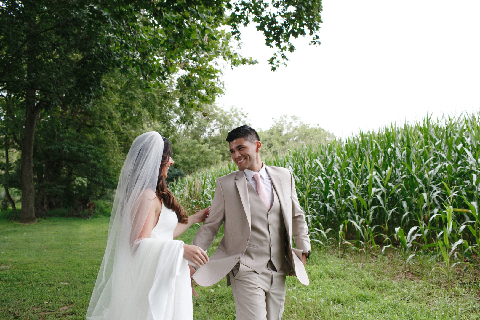 A bride and groom stroll through a grassy field bordering a cornfield, framed by trees during their enchanting Hotel Du Village wedding. The bride is radiant in her white dress and veil, while the groom looks dapper in his light-colored suit and tie.