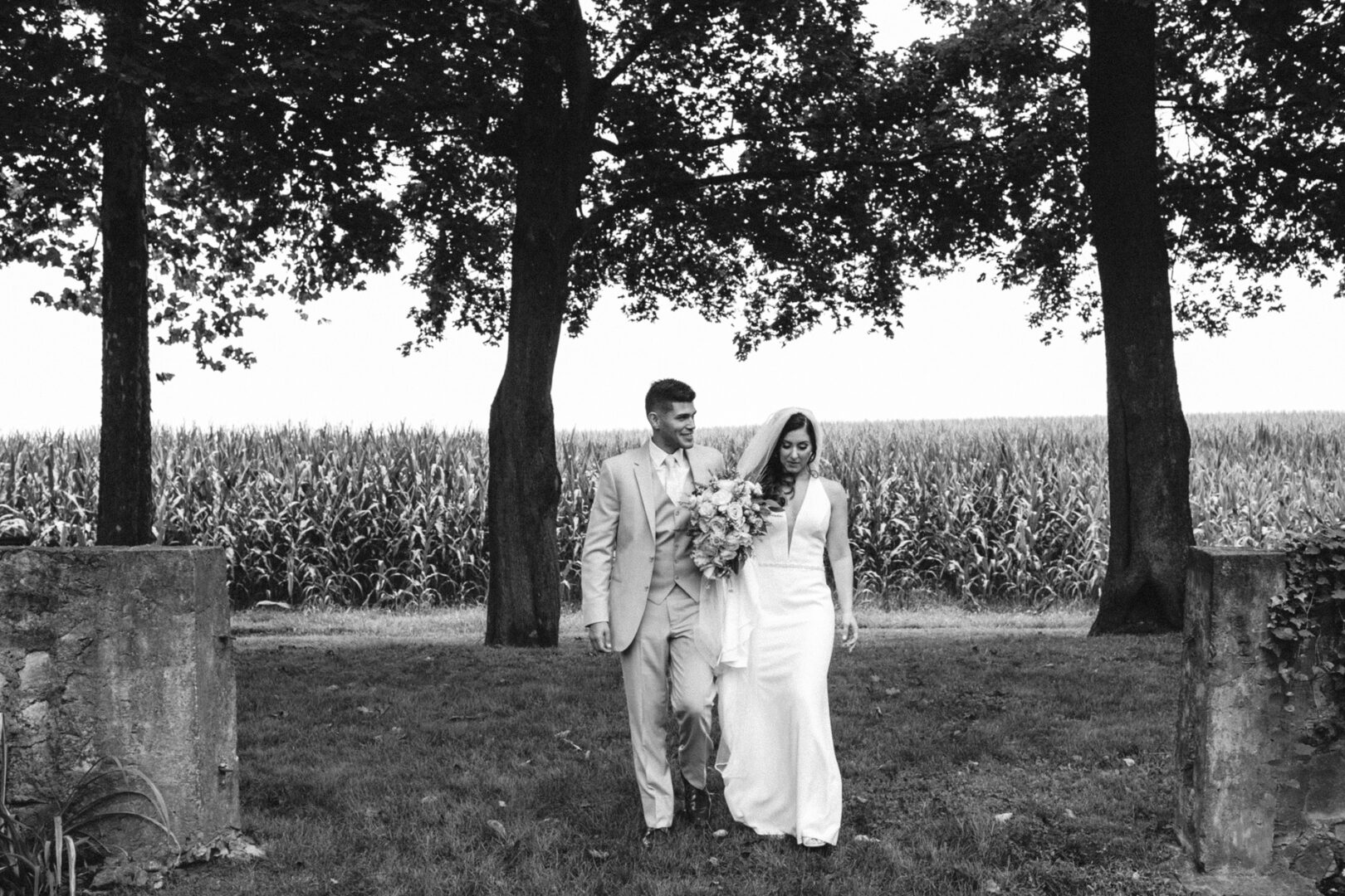 A bride and groom walk hand in hand through a grassy area at Hotel Du Village, surrounded by trees and a cornfield. The bride clutches her bouquet. Black and white image.