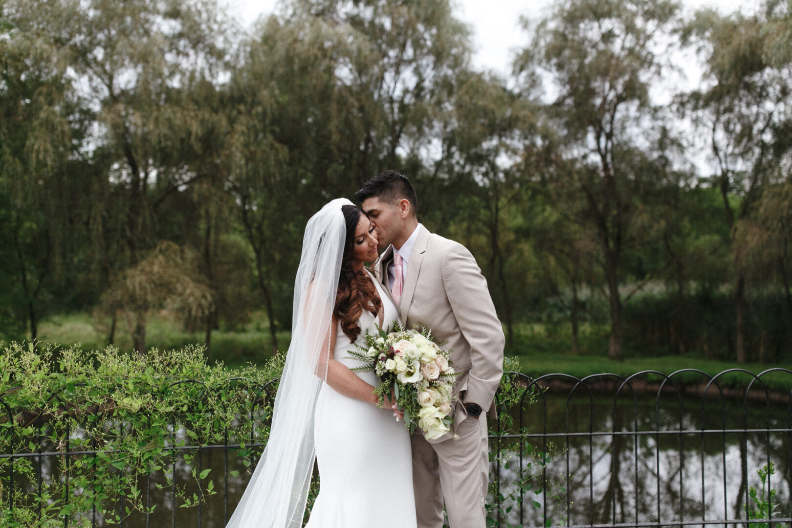 A bride and groom stand together outdoors by a lake at their enchanting Hotel Du Village wedding. The bride holds a bouquet, her long veil flowing, while the groom is in a light-colored suit, with trees softly framing the picturesque scene.