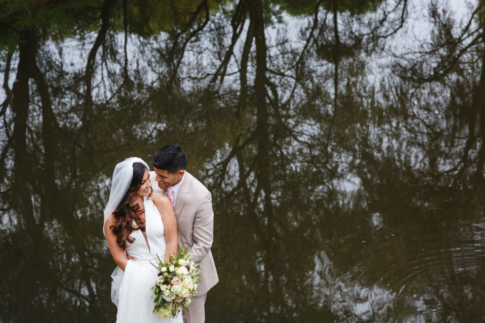 A couple in wedding attire poses by a pond at Hotel Du Village, their reflections mirrored in the water. The bride, clutching a bouquet, beams as the groom stands behind her. Graceful trees frame this romantic scene in the tranquil setting.