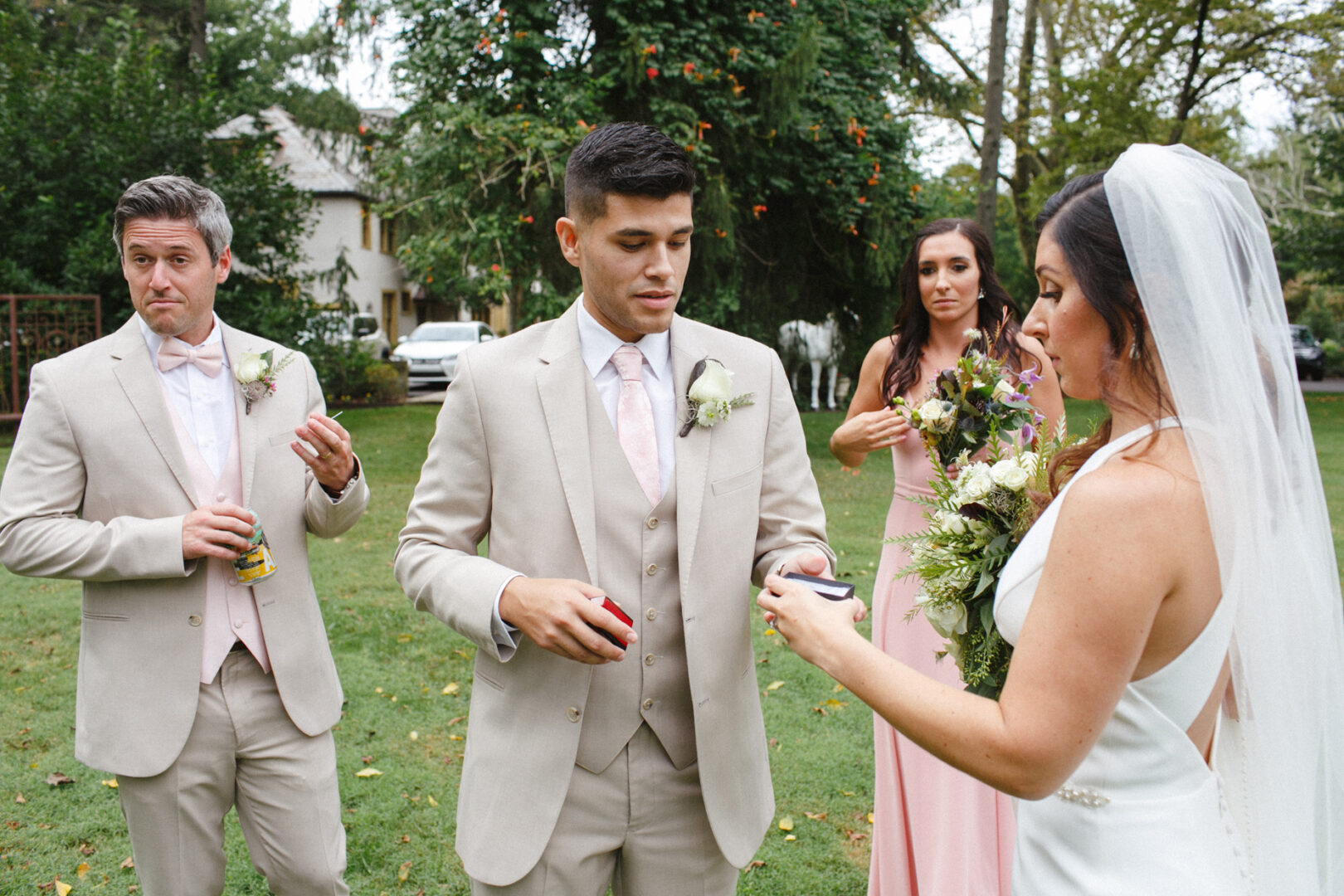 Bride and three others in formal attire stand outside at Hotel Du Village, holding items. One man has a drink, another a box, and a woman holds flowers. Lush greenery and the charming house enhance the backdrop of this picturesque wedding scene.