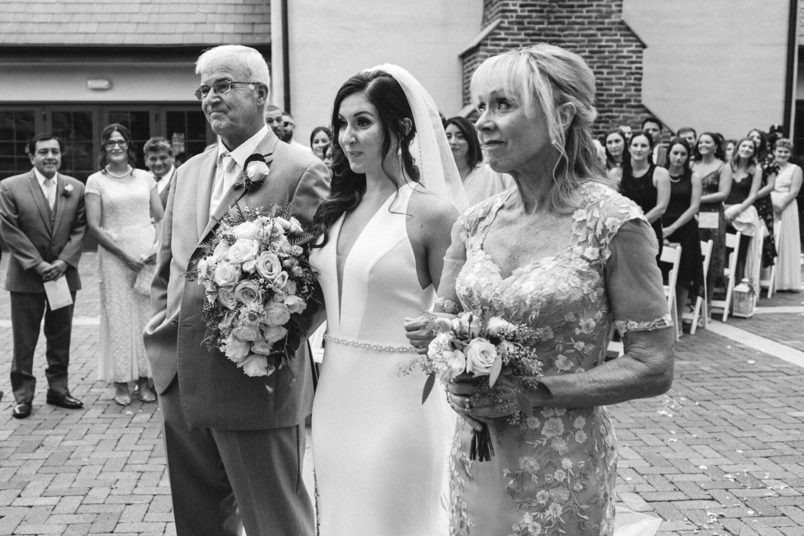 Bride in wedding dress holding flowers, standing between an older man in a suit and an older woman in a lace dress, with a crowd of people seated in the background. This black and white photo captures the timeless elegance of a Hotel Du Village wedding.