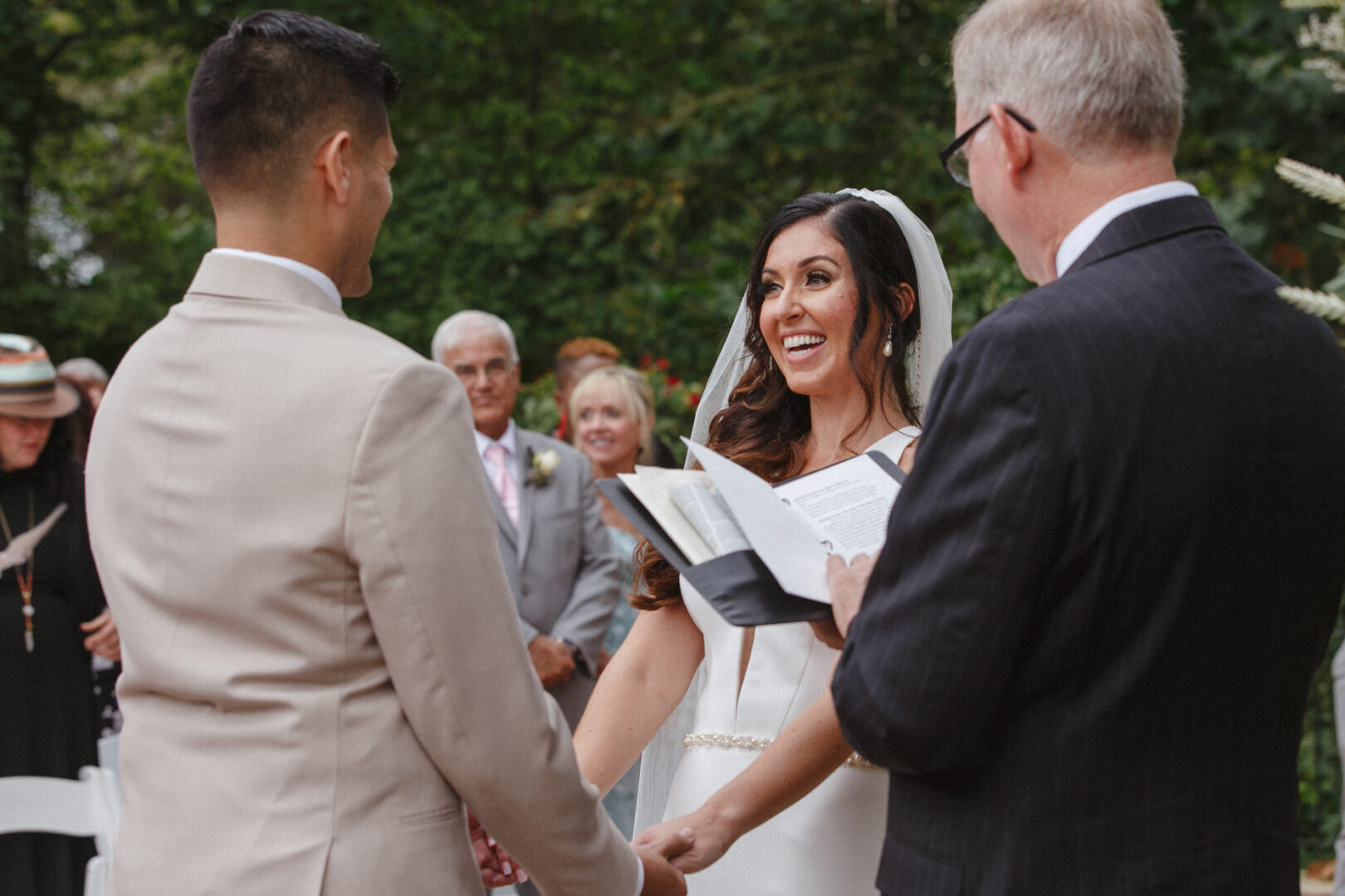 The bride and groom beam as they hold hands during their enchanting Hotel Du Village wedding, surrounded by an officiant and guests in the picturesque outdoor setting.