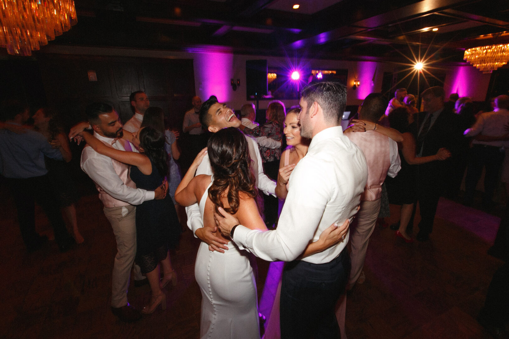 A group of people in formal attire are dancing closely on a dimly lit dance floor with purple lighting at a Hotel Du Village wedding. Other couples can be seen swaying gracefully in the background.