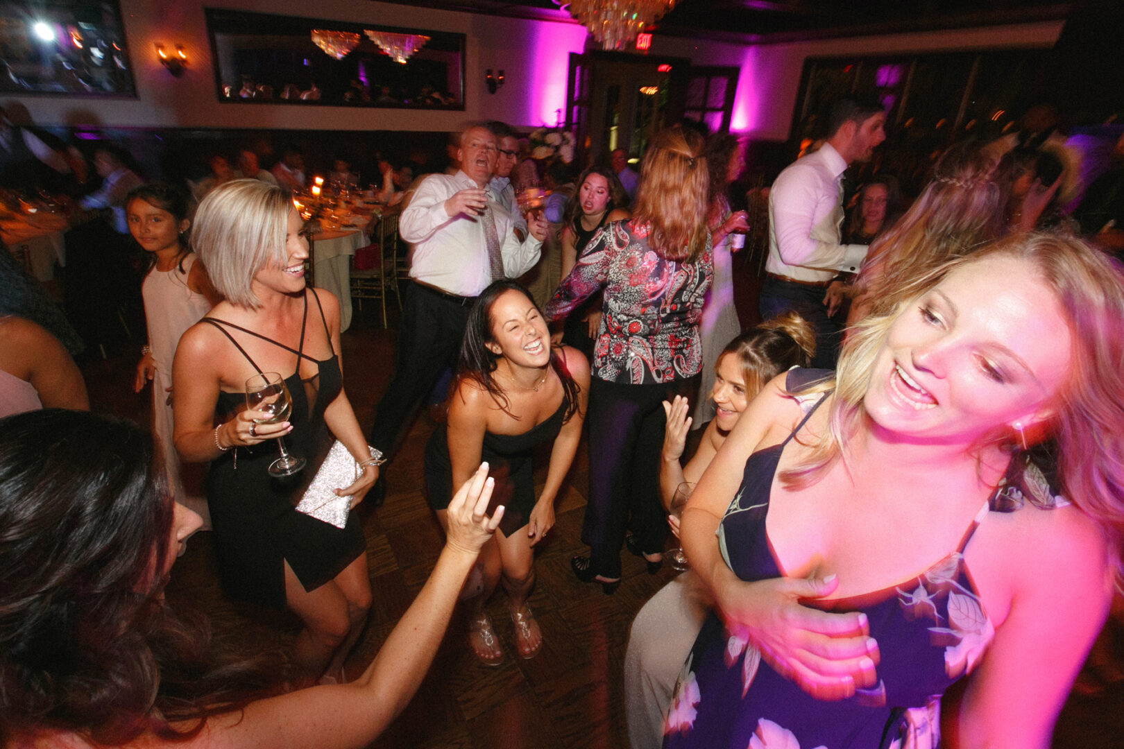 Amidst the colorful lights of a lively indoor party, guests joyfully dance and mingle, reminiscent of a Hotel Du Village Wedding celebration. A blend of standing and seated friends adds to the festive atmosphere.
