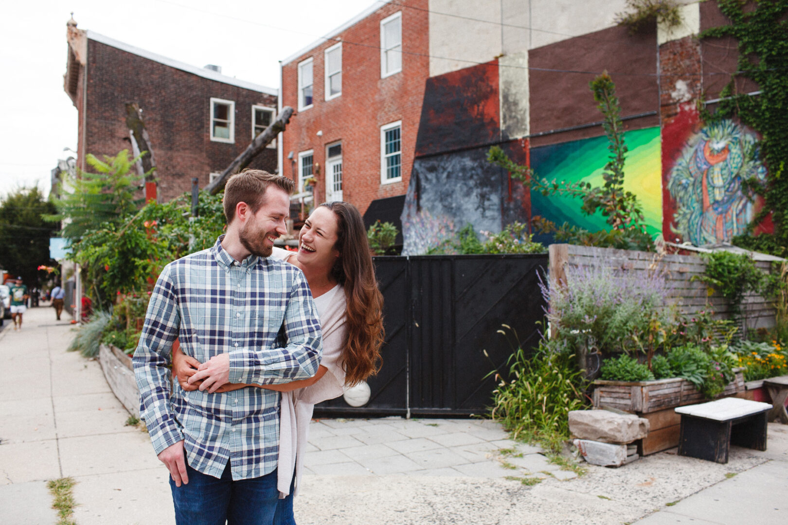 In the heart of Northern Liberties, a couple embraces and smiles amidst an urban setting adorned with vibrant murals and lush greenery, capturing the essence of their engagement session.