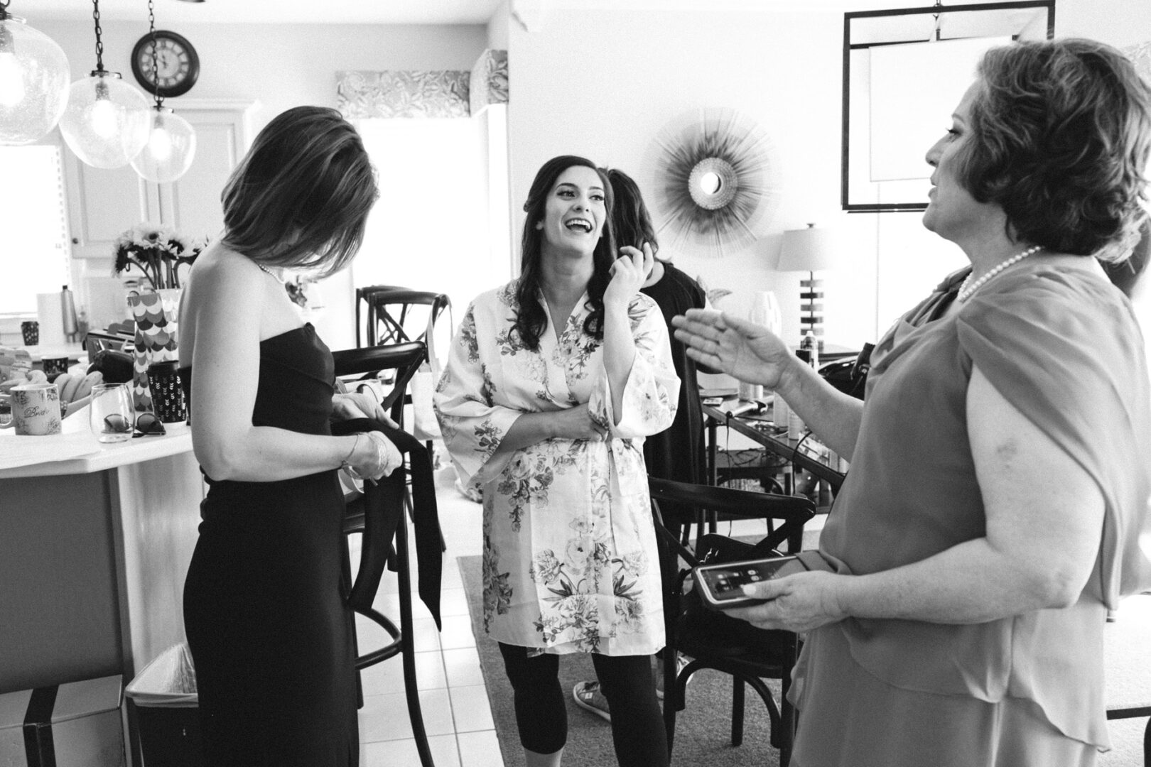 Three women are standing and talking in a kitchen area, reminiscent of an intimate gathering before a Stone Harbor Yacht Club wedding. One is wearing a floral robe, the others in dresses. They appear to be engaged in conversation, with one woman holding a phone.