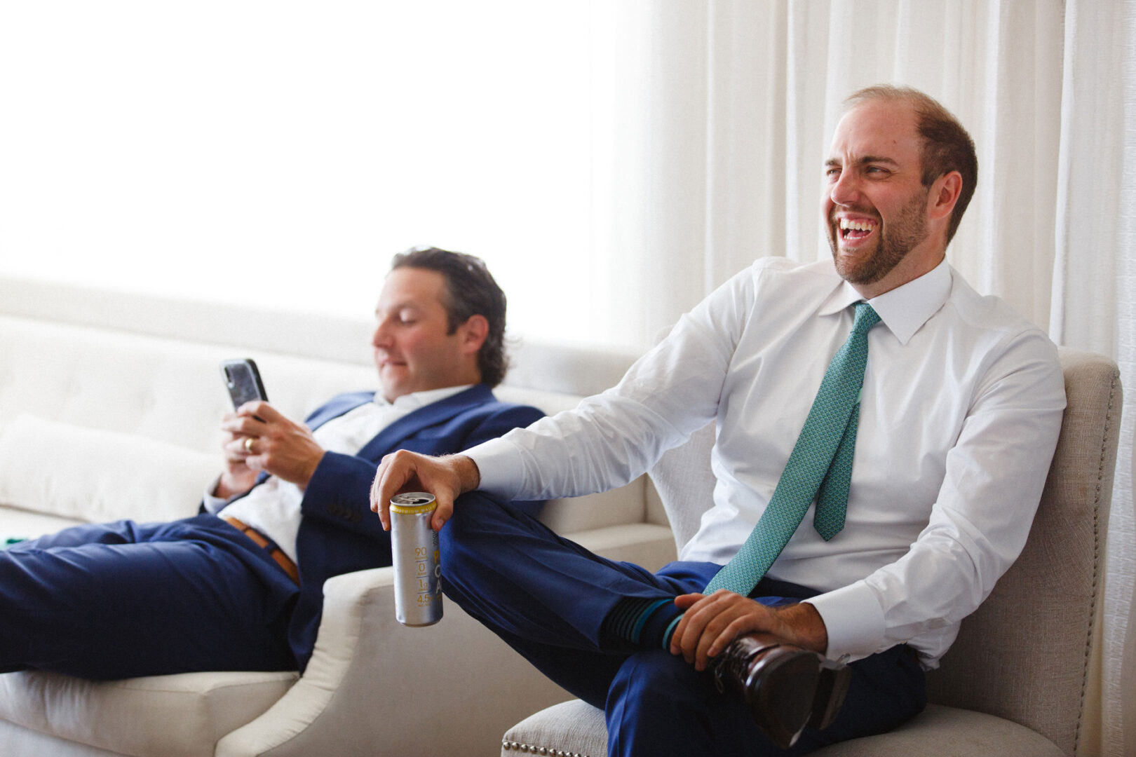In a bright room with white curtains, two men in suits sit on chairs at the Stone Harbor Yacht Club Wedding. One man, holding a can, bursts into laughter while the other focuses intently on his phone.