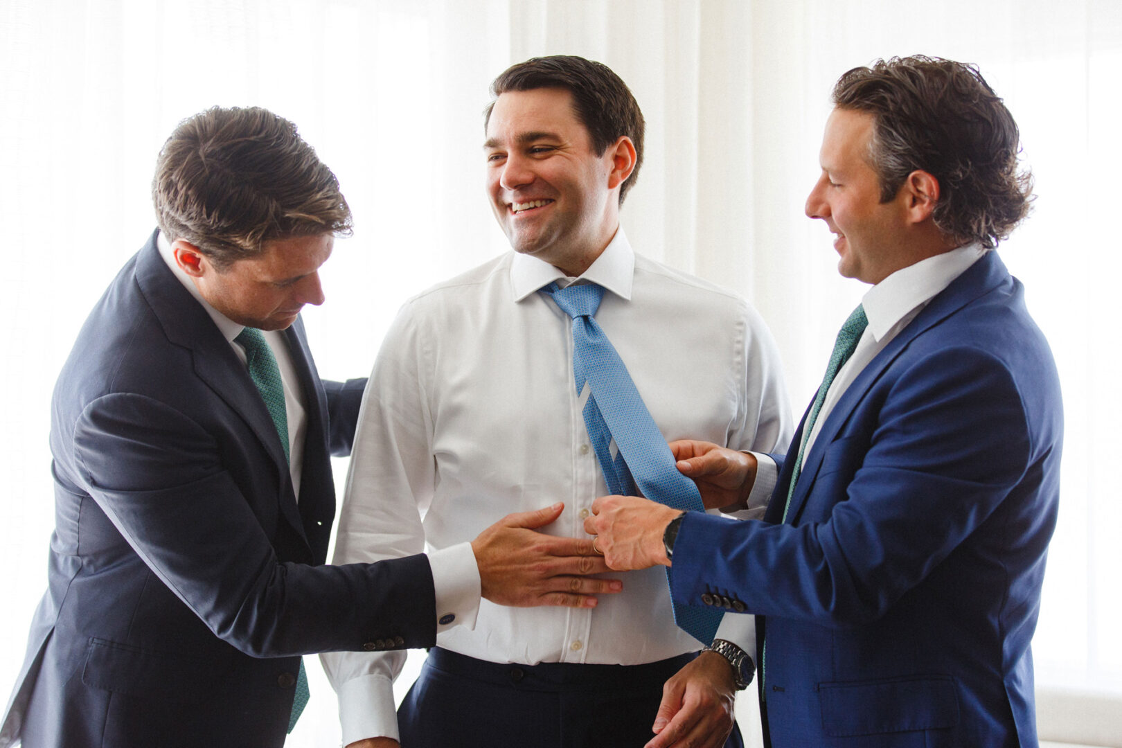 Three men in suits, two adjusting the tie of the smiling one in the middle, as if preparing for a Stone Harbor Yacht Club Wedding. They are in a brightly lit room.
