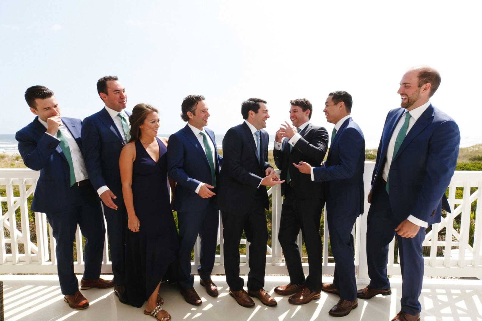 A group of seven men and one woman in formal attire, standing on a balcony at the Stone Harbor Yacht Club wedding. They're laughing and engaged in conversation, framed by a sunny outdoor background.