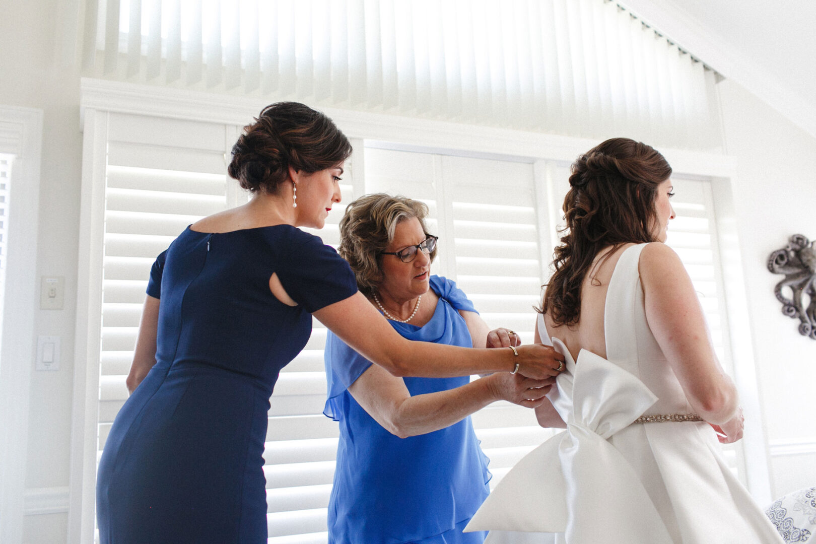 Three women in a room are helping one woman adjust her white dress with a large bow at the back, preparing for the elegance of a Stone Harbor Yacht Club wedding. They are indoors with white blinds in the background.
