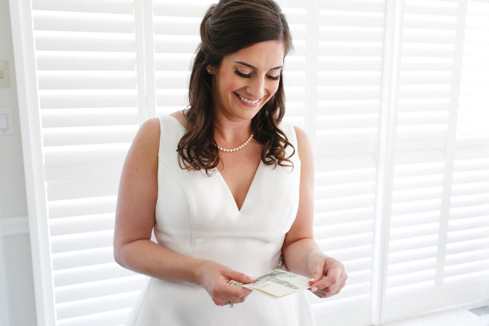 A woman in a white dress stands indoors at the Stone Harbor Yacht Club Wedding, smiling while holding papers. White shutters frame the background.