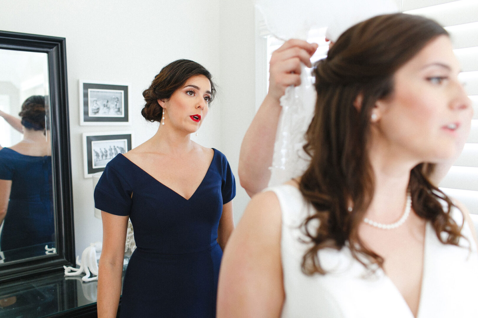 At a Stone Harbor Yacht Club wedding, a woman in a blue dress stands near another in a white dress, who is having her hair arranged. They are indoors with framed pictures on the wall, capturing the joyous ambiance of the day.