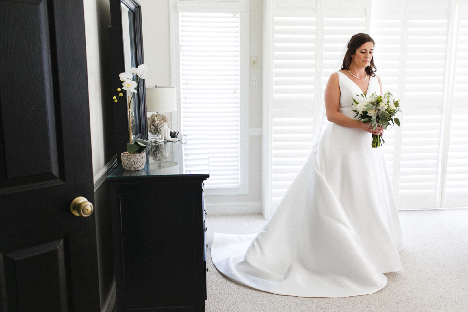 A bride in a long white gown holds a bouquet of flowers, standing in a sunlit room with white shutters and a black dresser, capturing the elegance of her Stone Harbor Yacht Club wedding.