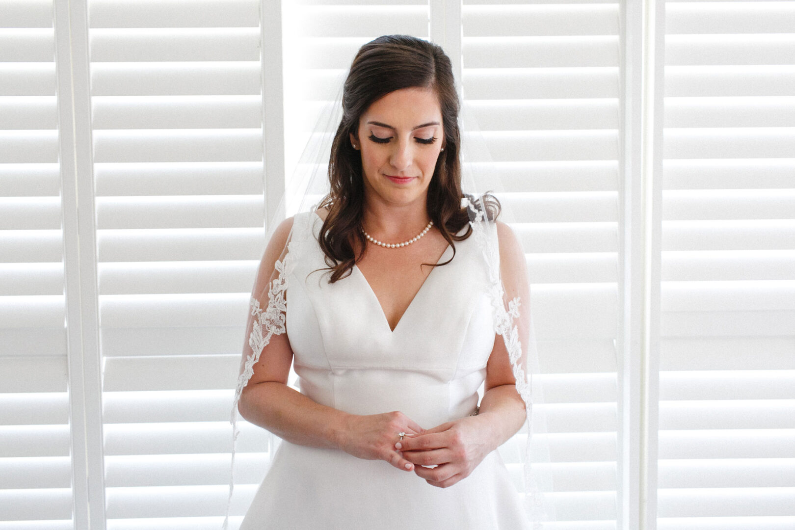 A bride in a white dress stands gracefully in front of white shutters at the Stone Harbor Yacht Club, looking down. She wears a pearl necklace, with delicate lace sleeves and a flowing veil that adds to her timeless elegance.