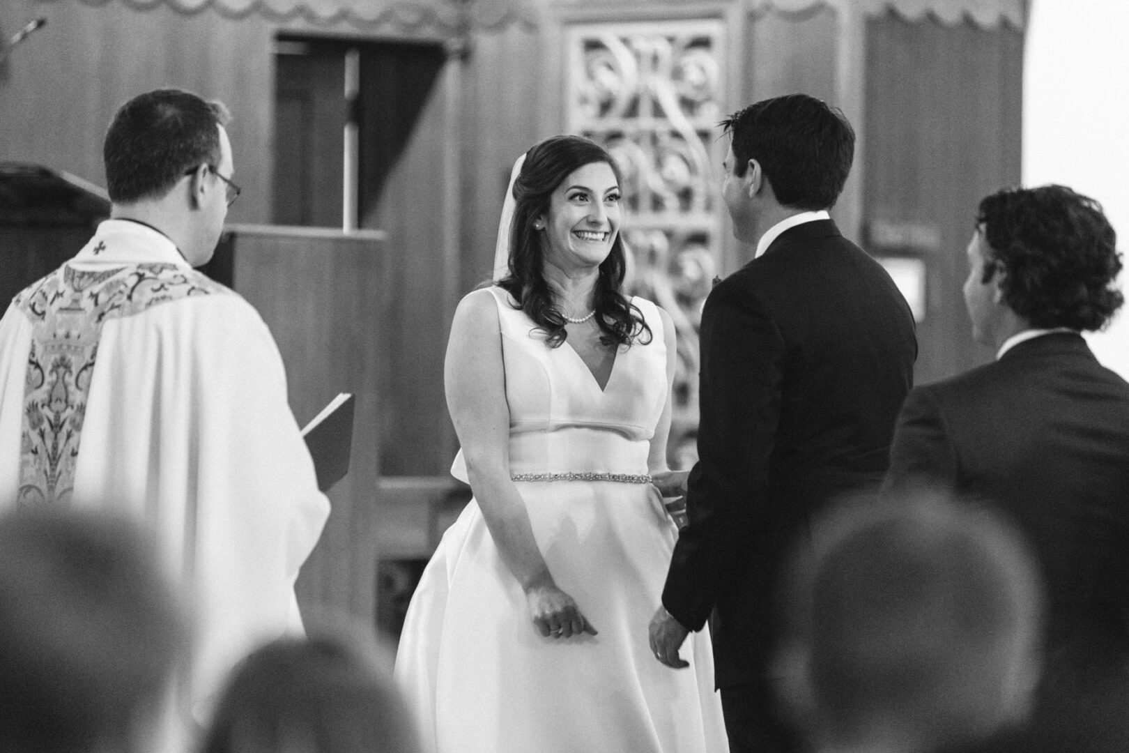 A bride and groom exchange smiles at their Stone Harbor Yacht Club Wedding ceremony, with a person in clerical attire standing nearby.