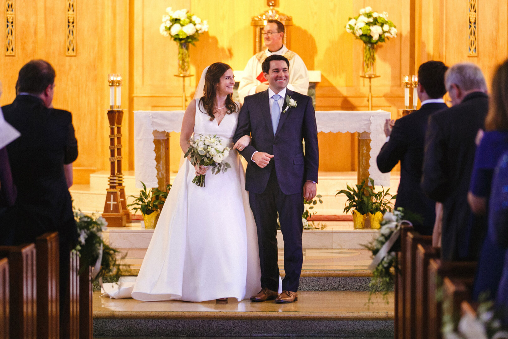 A bride and groom stand together at the altar, smiling and holding flowers at their charming Stone Harbor Yacht Club wedding. The priest stands behind them as guests are seated on either side, creating a picturesque ceremony setting.