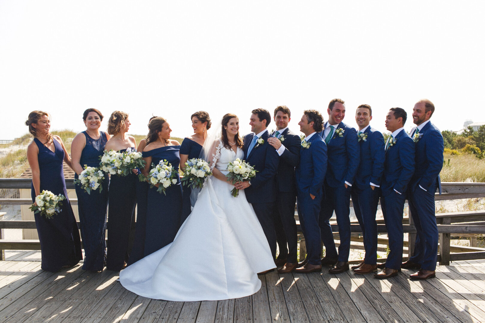 At the Stone Harbor Yacht Club wedding, a joyful party gathers on the wooden deck. The bride, radiant in her white gown, holds a bouquet as she's surrounded by bridesmaids in navy dresses and groomsmen in blue suits, all capturing the moment with bright smiles.