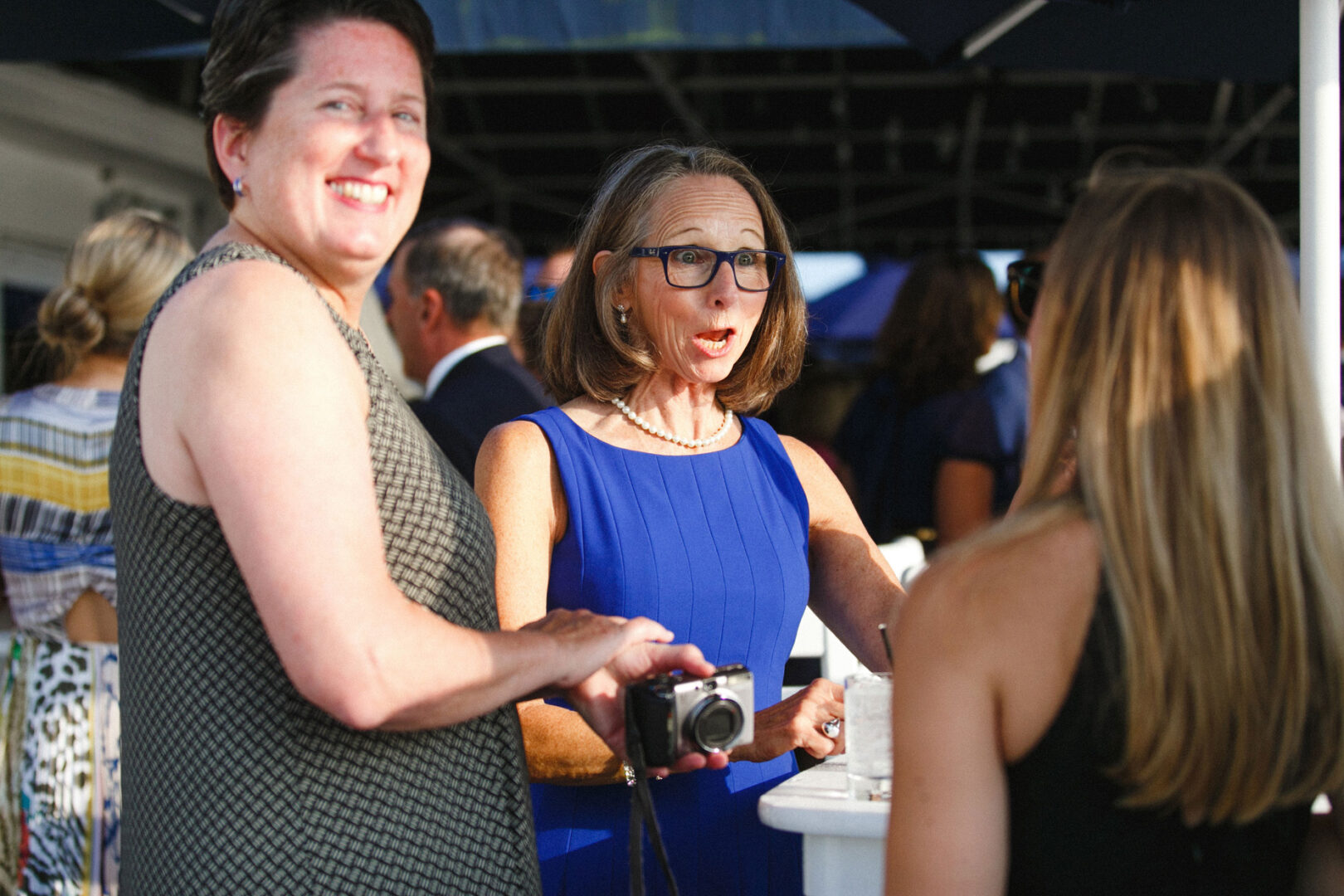 Two women are smiling and chatting with another woman at a vibrant outdoor gathering, perhaps capturing special moments at a Stone Harbor Yacht Club wedding. One holds a camera while the other gestures animatedly, encapsulating the joy of the occasion.